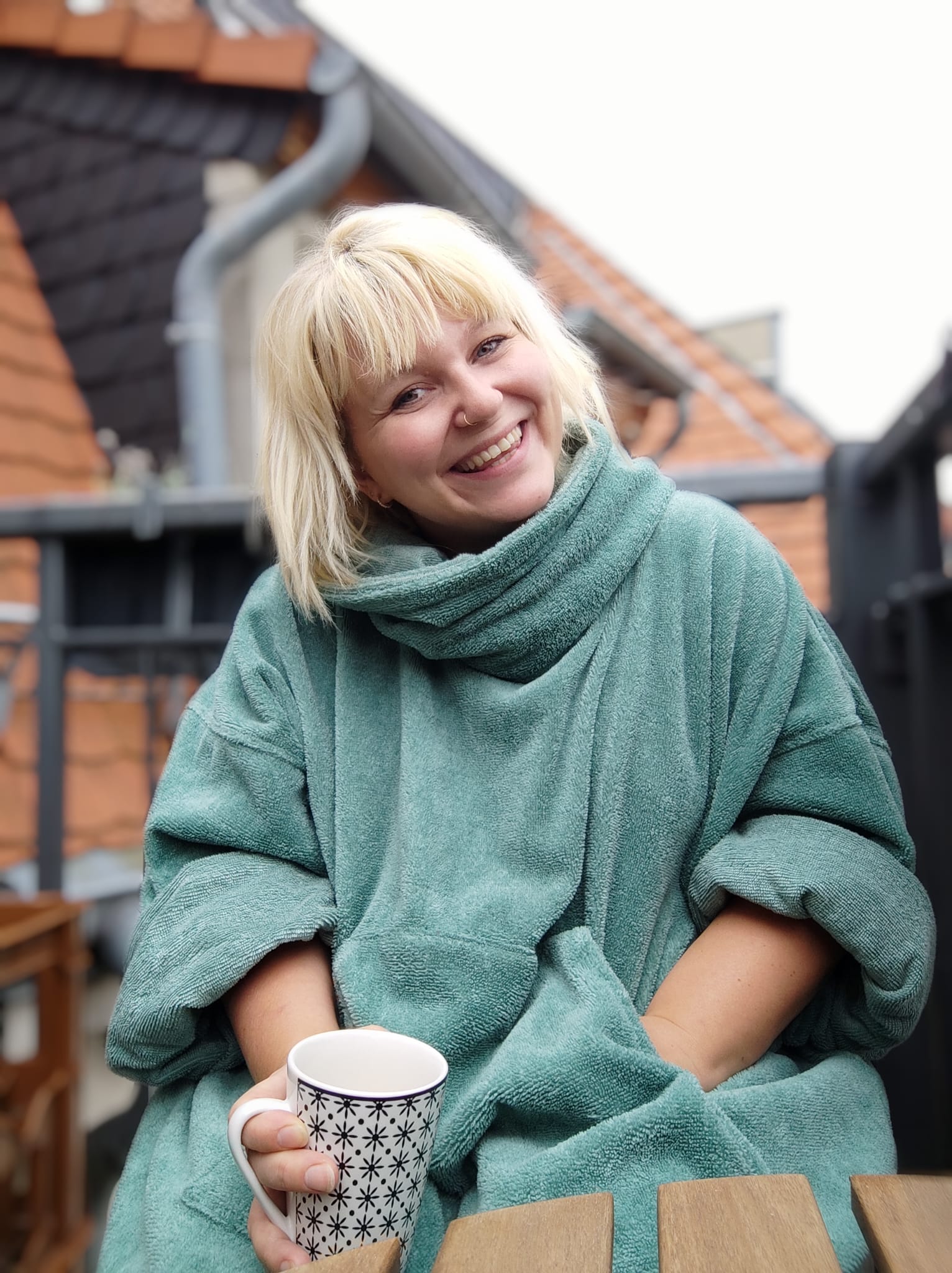 Blonde lächelnde Frau trägt Badeponcho in Mint und hält eine Tasse in der Hand